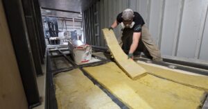 A worker wearing a safety gas mask installing insulation foam into storage building on a construction site.