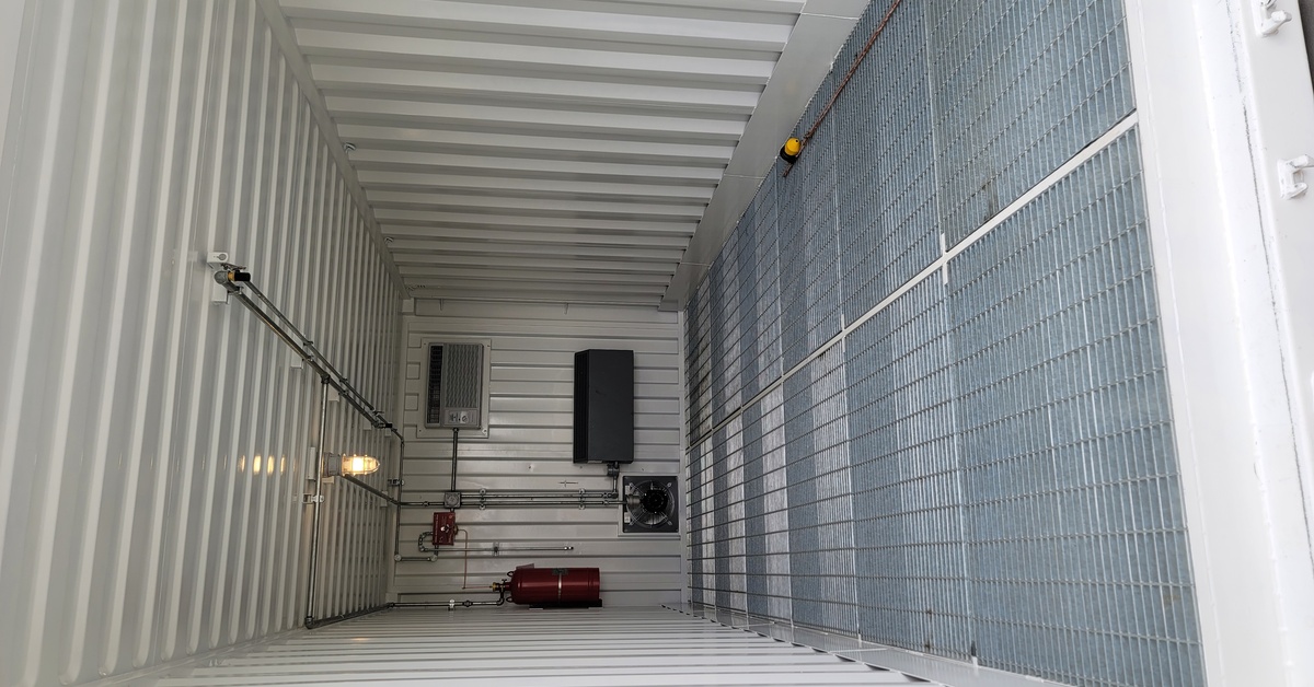 The interior of a large, white storage container. Several pipes and boxes are attached to the back wall of the container.