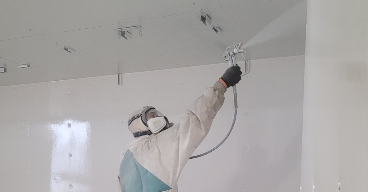 A man in full safety gear and a mask spraying paint onto the inside of a shipping container with a paint gun.