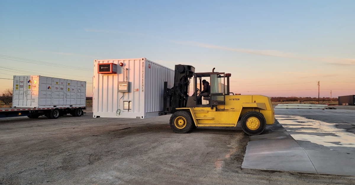 A large, yellow forklift lifting a storage container off of the ground. It is moving it towards a truck in the background.