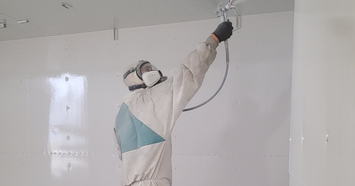 A man wearing full protective gear spraying a protective coating on the inside of a white storage container.