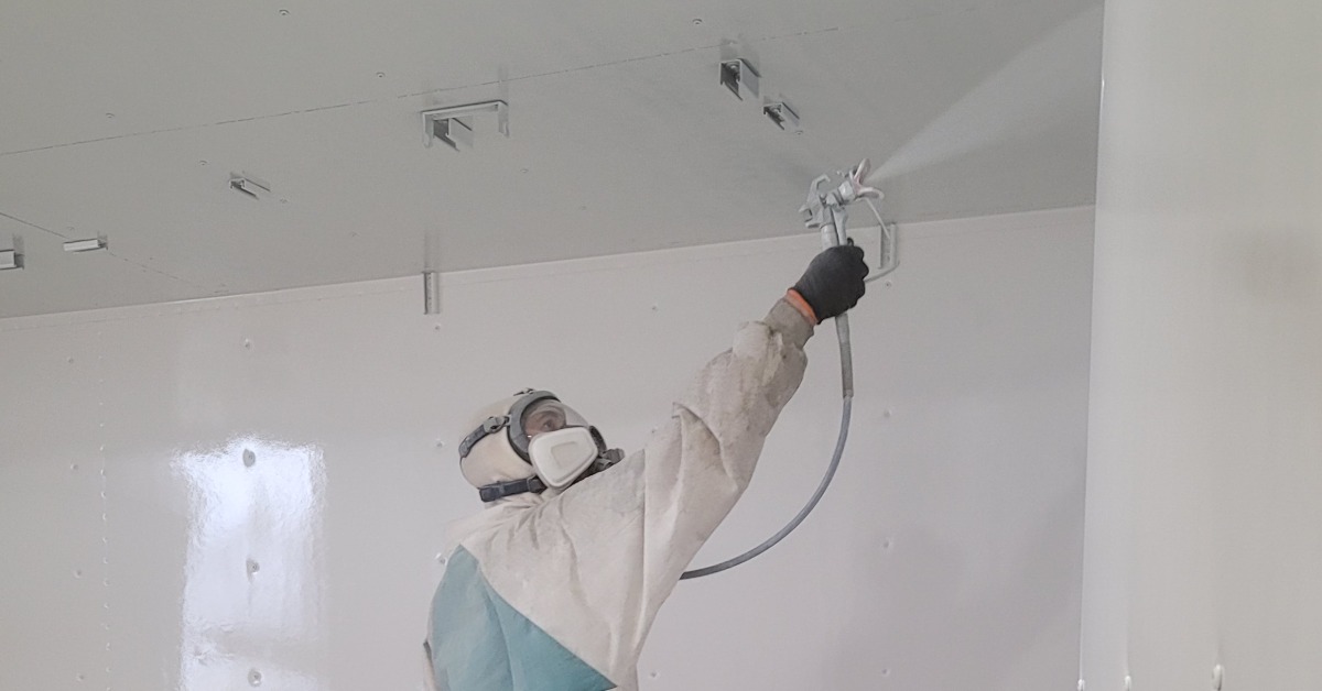 A man in white protective gear and a mask praying the inside of a storage container with a white paint.