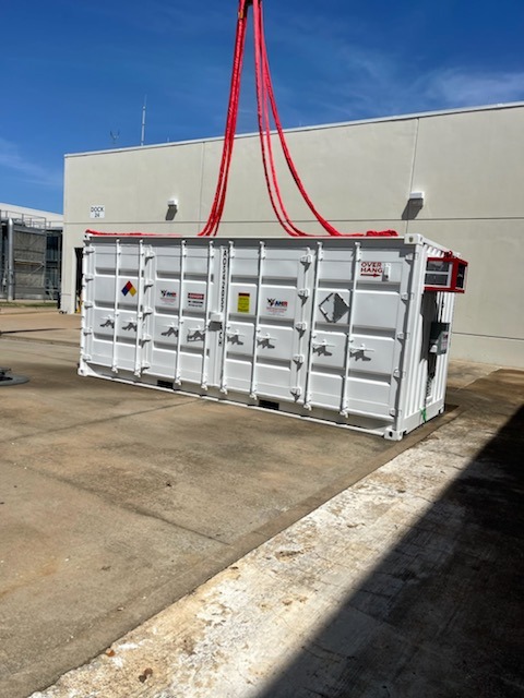 A white storage container sitting in a docking area by itself. There are large, red cables attached to the top of it.